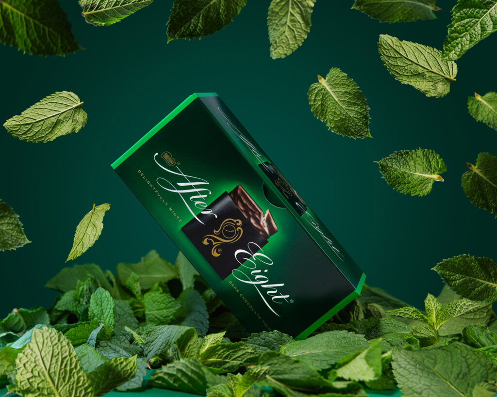 After Eight dark chocolate thins box in deep green packaging, surrounded by fresh, flying mint leaves and resting on a bed of mint, against a dark green background.