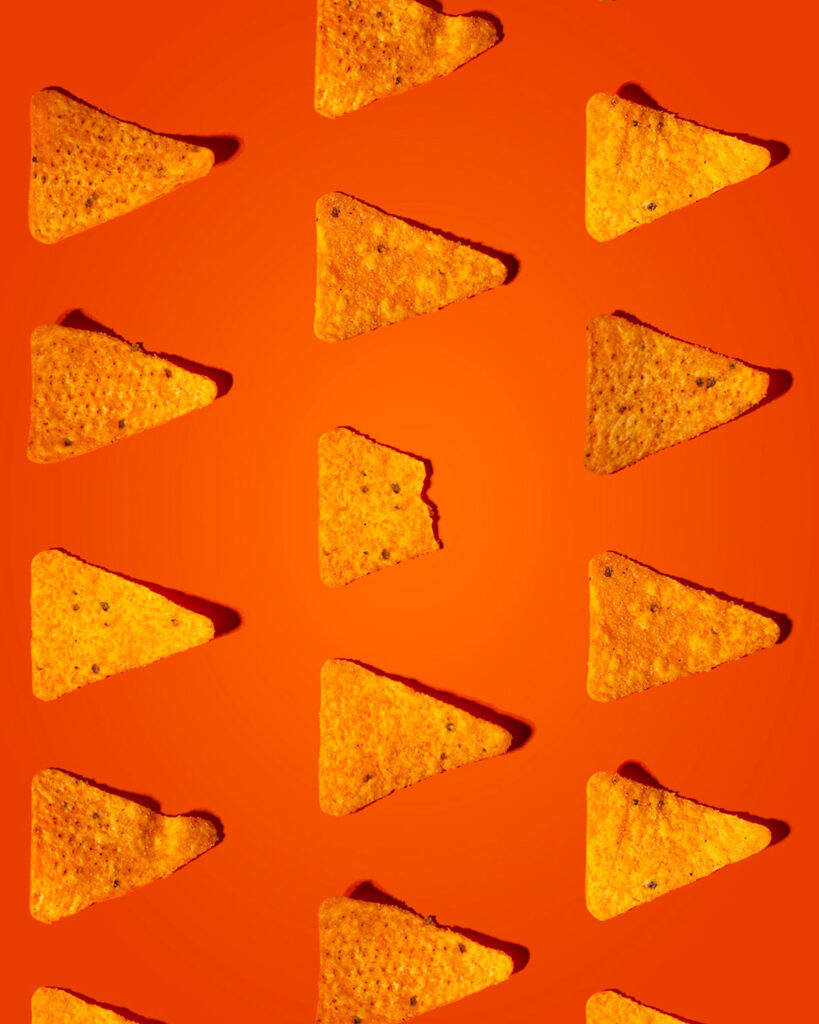 Repeating pattern of crispy triangular tortilla chips (nachos) on a vibrant orange background, with one central chip featuring a small bite taken out.