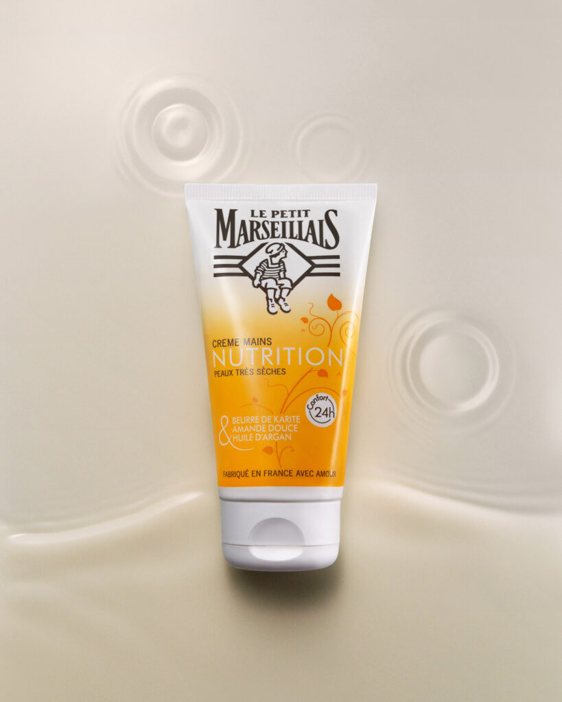 Le Petit Marseillais Crème Mains Nutrition hand cream tube centered in a creamy, beige liquid splash or ripple effect, emphasizing the moisturizing texture.