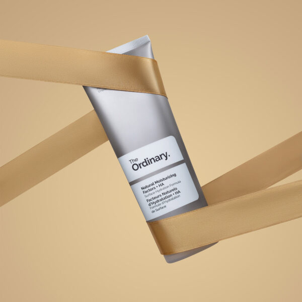 The Ordinary Natural Moisturizing Factors + HA cream tube, wrapped diagonally with a flowing gold ribbon against a beige background, emphasizing a gift or luxury feel.