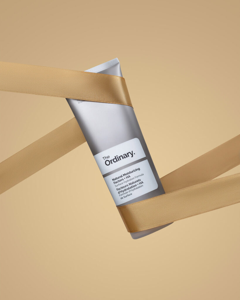 The Ordinary Natural Moisturizing Factors + HA cream tube, wrapped diagonally with a flowing gold ribbon against a beige background, emphasizing a gift or luxury feel.