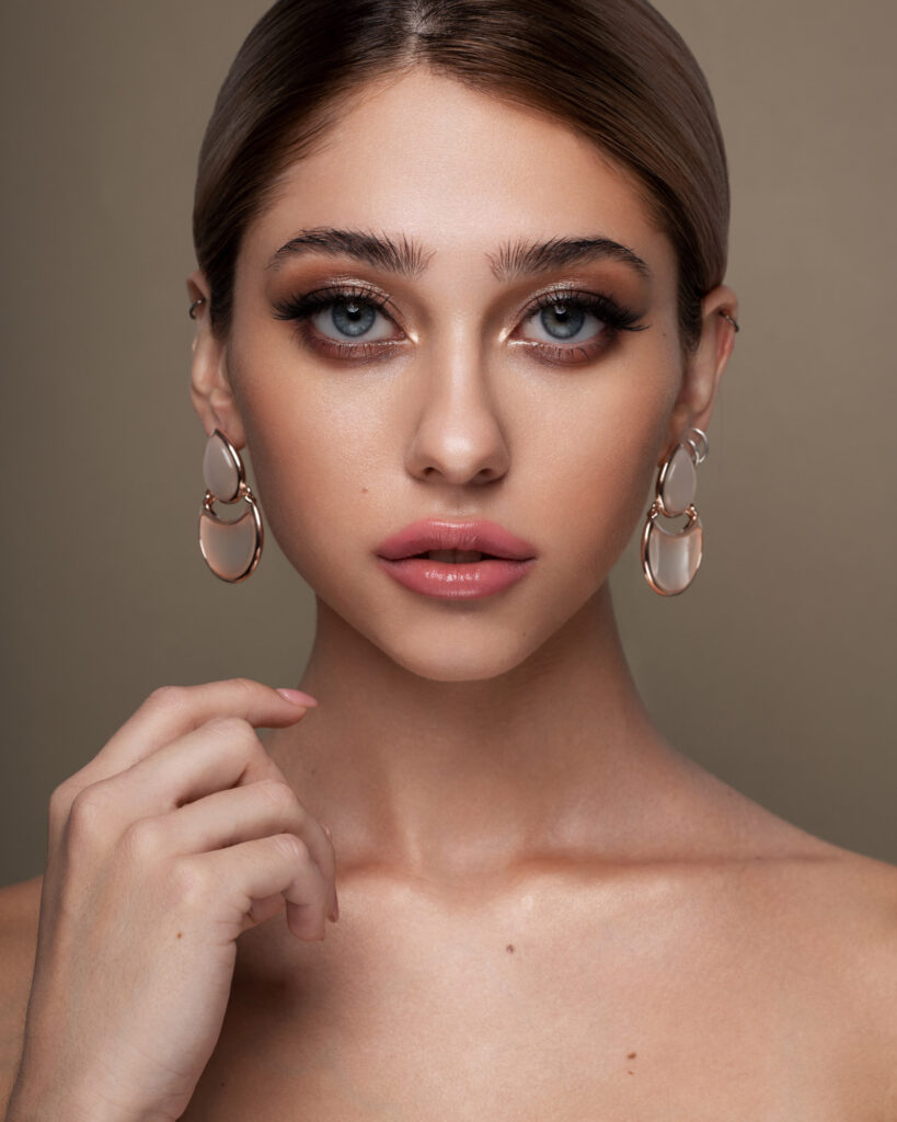 High-end retouched glamour portrait with dramatic eye makeup, glossy pink lips, and large rose gold statement earrings, showcasing flawless skin and perfect hair detail