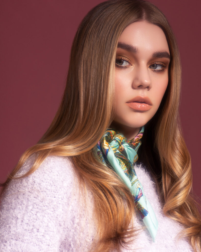 High-end retouched fashion portrait of a model with long blonde wavy hair, wearing a fuzzy lavender sweater and a silk patterned scarf.