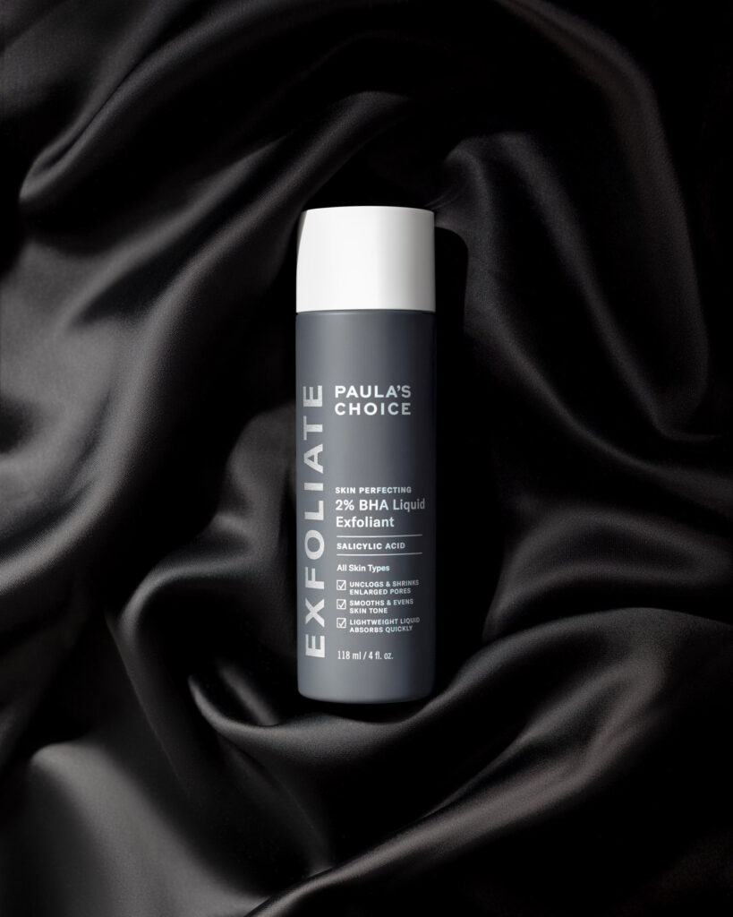 Paula's Choice Skin Perfecting 2% BHA Liquid Exfoliant bottle centered on a luxurious, draped black satin or silk fabric, conveying a sense of sophistication and smoothness.
