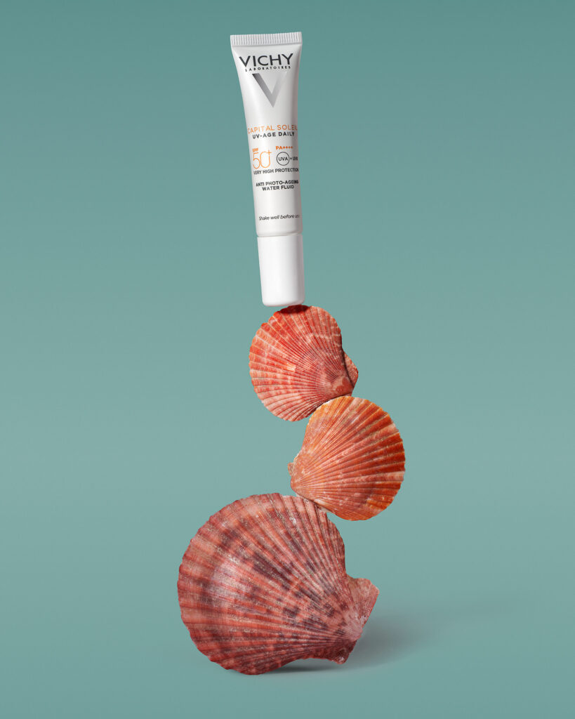 Vichy Capital Soleil UV-Age Daily SPF 50+ sunscreen tube balanced on a stack of three natural reddish-orange seashells against a teal or sea-green background.