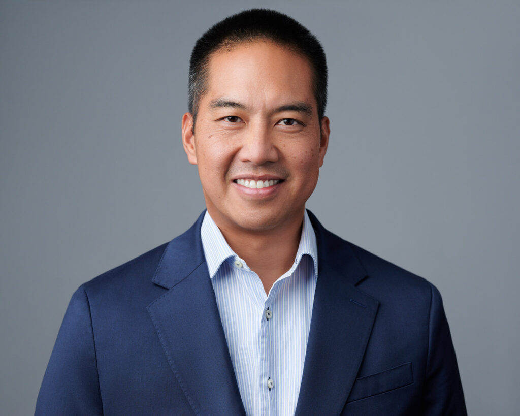 Smiling Asian male business professional in a navy blue suit and striped shirt, photographed against a neutral grey studio background for a corporate portrait.