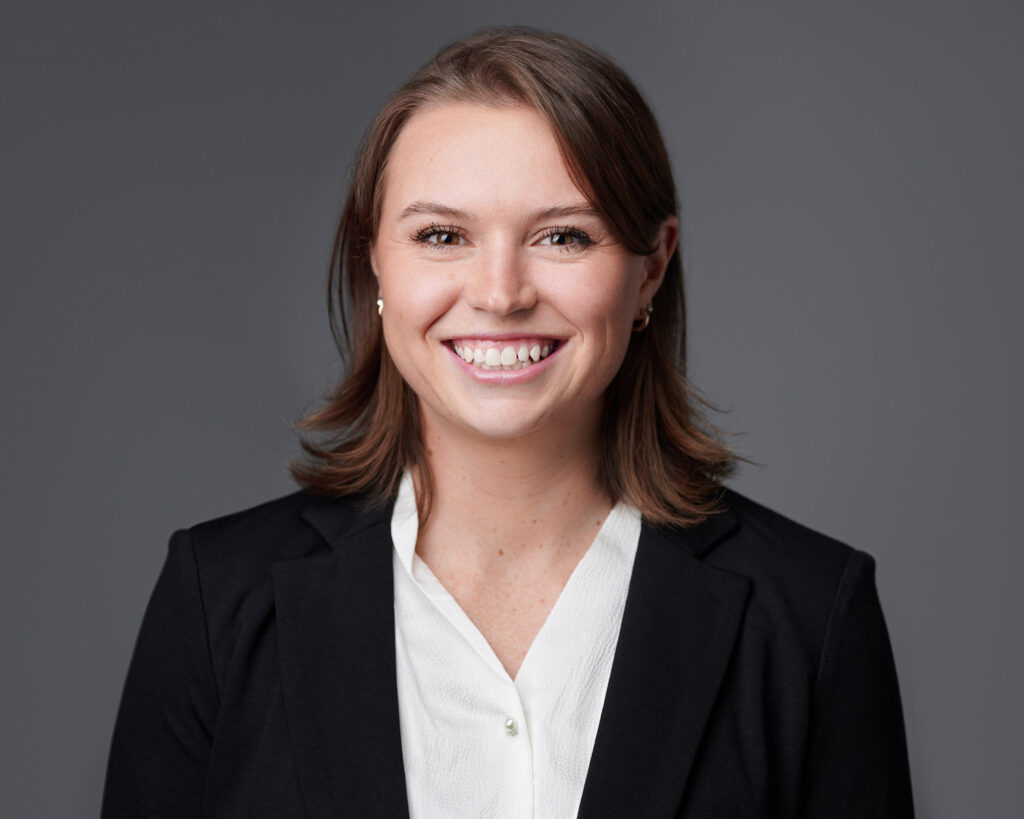 Smiling young female business professional in a black blazer and white blouse, photographed against a smooth grey studio background for a corporate headshot.
