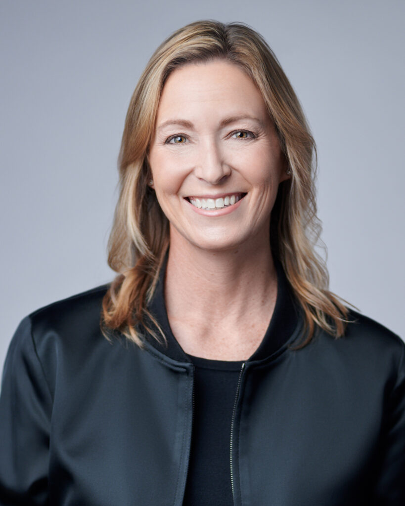 Smiling middle-aged female business professional in a black satin bomber jacket, photographed against a smooth, neutral grey studio background for a corporate headshot.