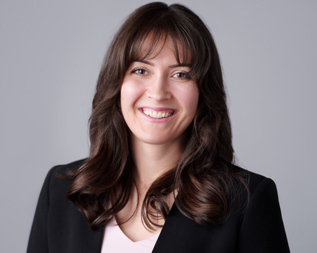 Smiling young female business professional with long dark hair, wearing a black blazer over a light pink top, photographed against a neutral grey studio background.