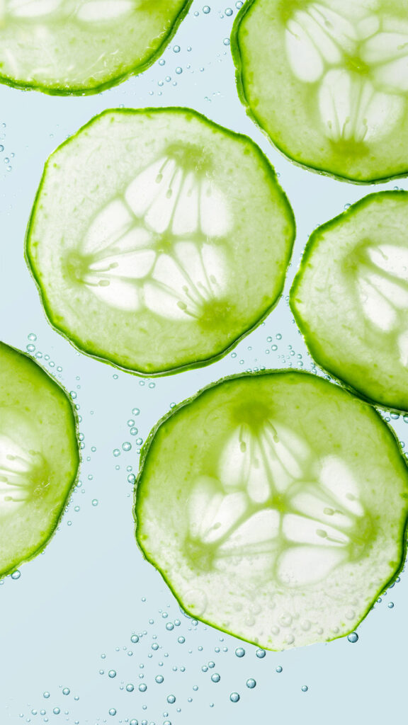 Extreme close-up macro shot of translucent fresh cucumber slices submerged in water with rising bubbles, suggesting a cooling and refreshing effect for skincare