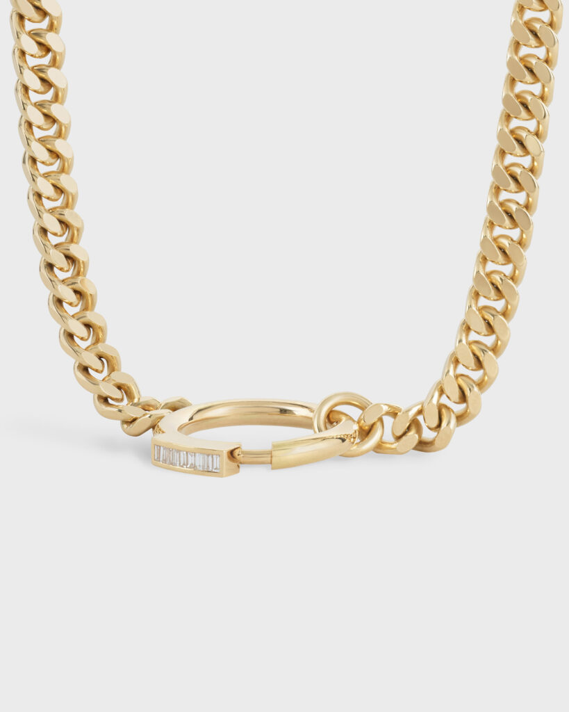 Close-up of a thick gold Cuban or curb link chain necklace with a textured, modern carabiner clasp paved with baguette diamonds, professionally retouched on a light background.
