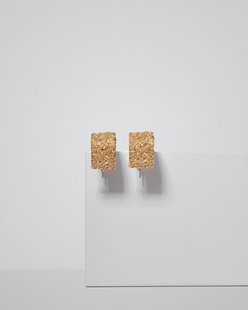 Pair of gold-plated square stud earrings with a rough, textured finish, suspended against a minimalist white background with subtle shadow casting.