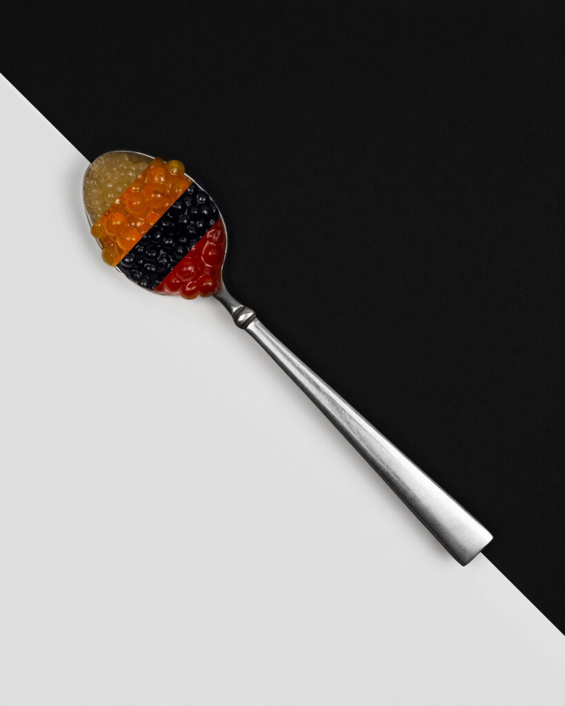 Silver spoon holding three stripes of yellow, orange, red, and black colored gourmet caviar, placed diagonally across a minimalist black and white split background.