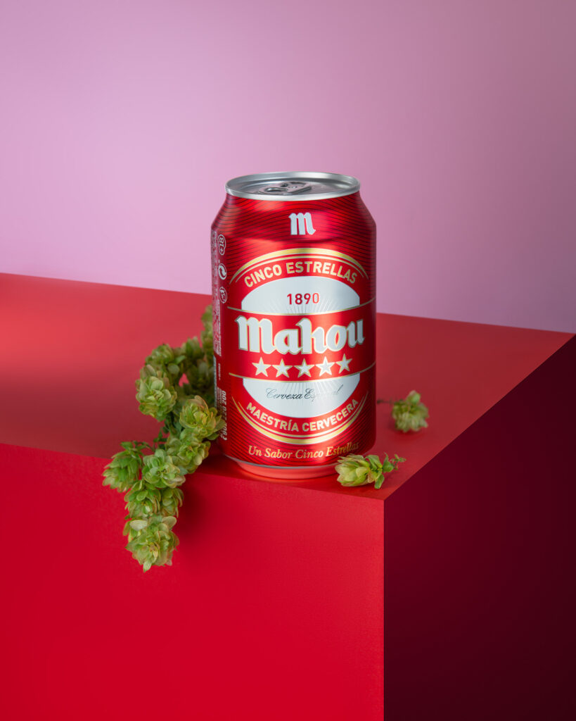 Mahou Cinco Estrellas red beer can placed on a bright red geometric prop, with green hop cones arranged next to it, against a pink and purple gradient background.