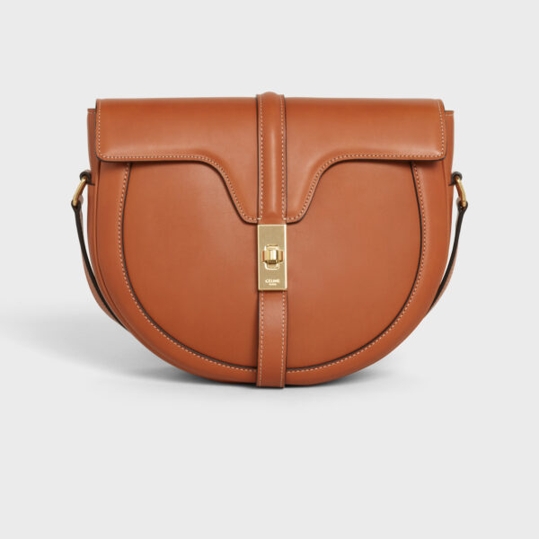 Luxury Celine tan/camel leather saddle bag with curved silhouette, central strap, and rectangular gold metal clasp, photographed for e-commerce on a light grey background.