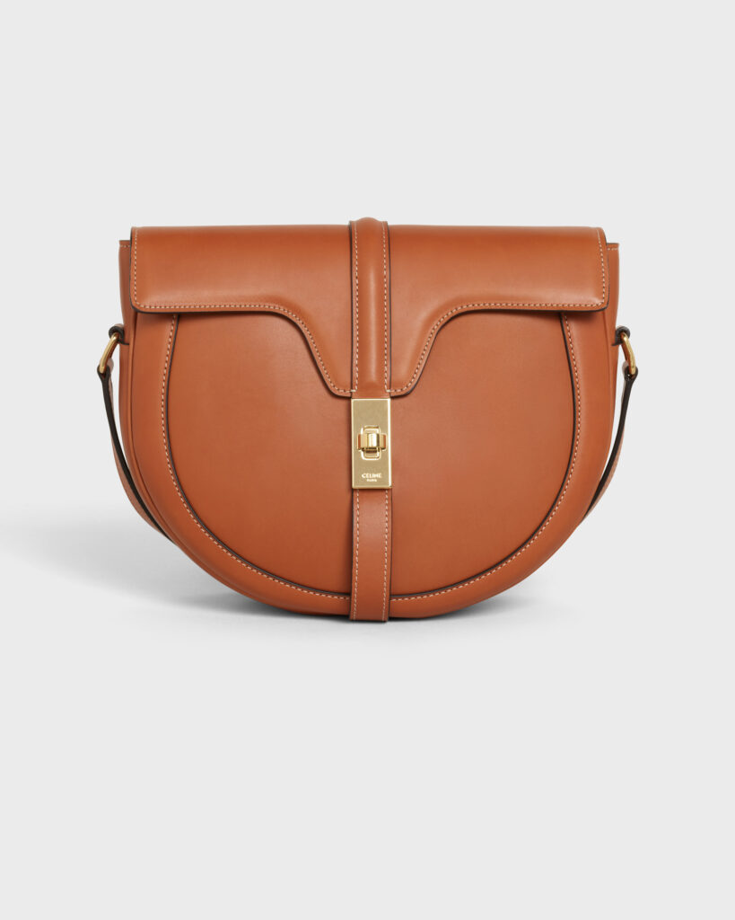 Luxury Celine tan/camel leather saddle bag with curved silhouette, central strap, and rectangular gold metal clasp, photographed for e-commerce on a light grey background.
