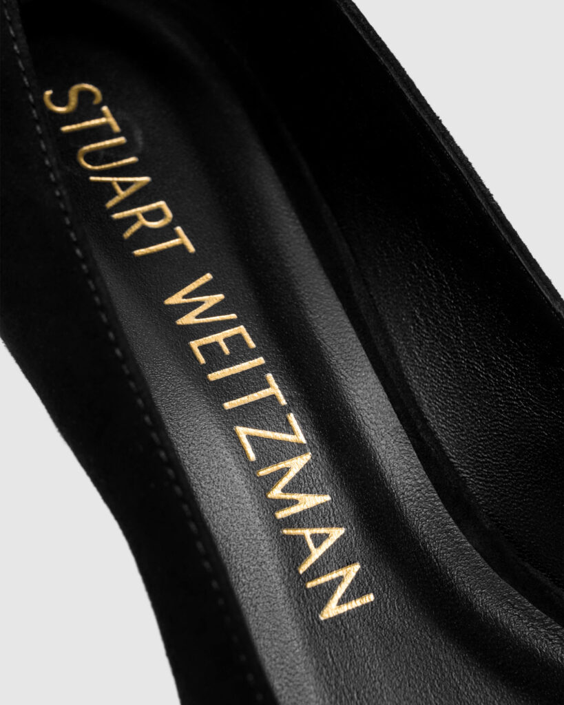 Extreme close-up macro shot of the Stuart Weitzman logo debossed in gold foil on a black leather insole, highlighting the luxury branding detail.