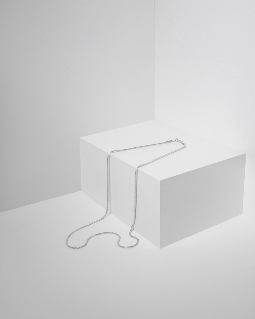 Elegant silver snake or box chain necklace draped over a white geometric display cube, photographed against a seamless white background in a high-key setting.